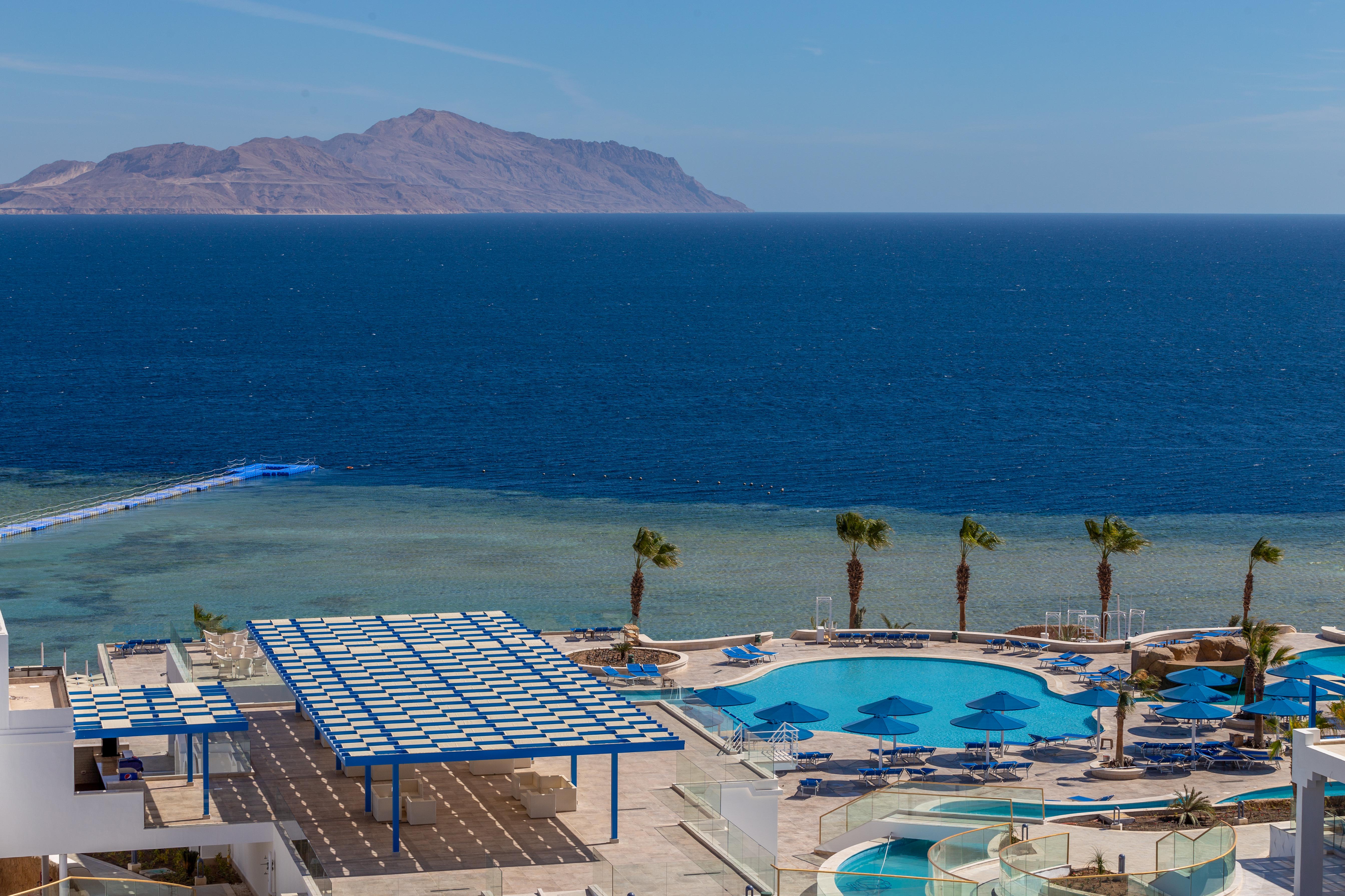 Pickalbatros Palace Sharm - "Aqua Park" Exterior foto The photo shows a beautiful coastal scene featuring a large swimming pool area with lounge chairs and umbrellas. In the background, there is a clear blue ocean stretching out to the horizon, with a distant landmass or mountain visible. Palm trees are