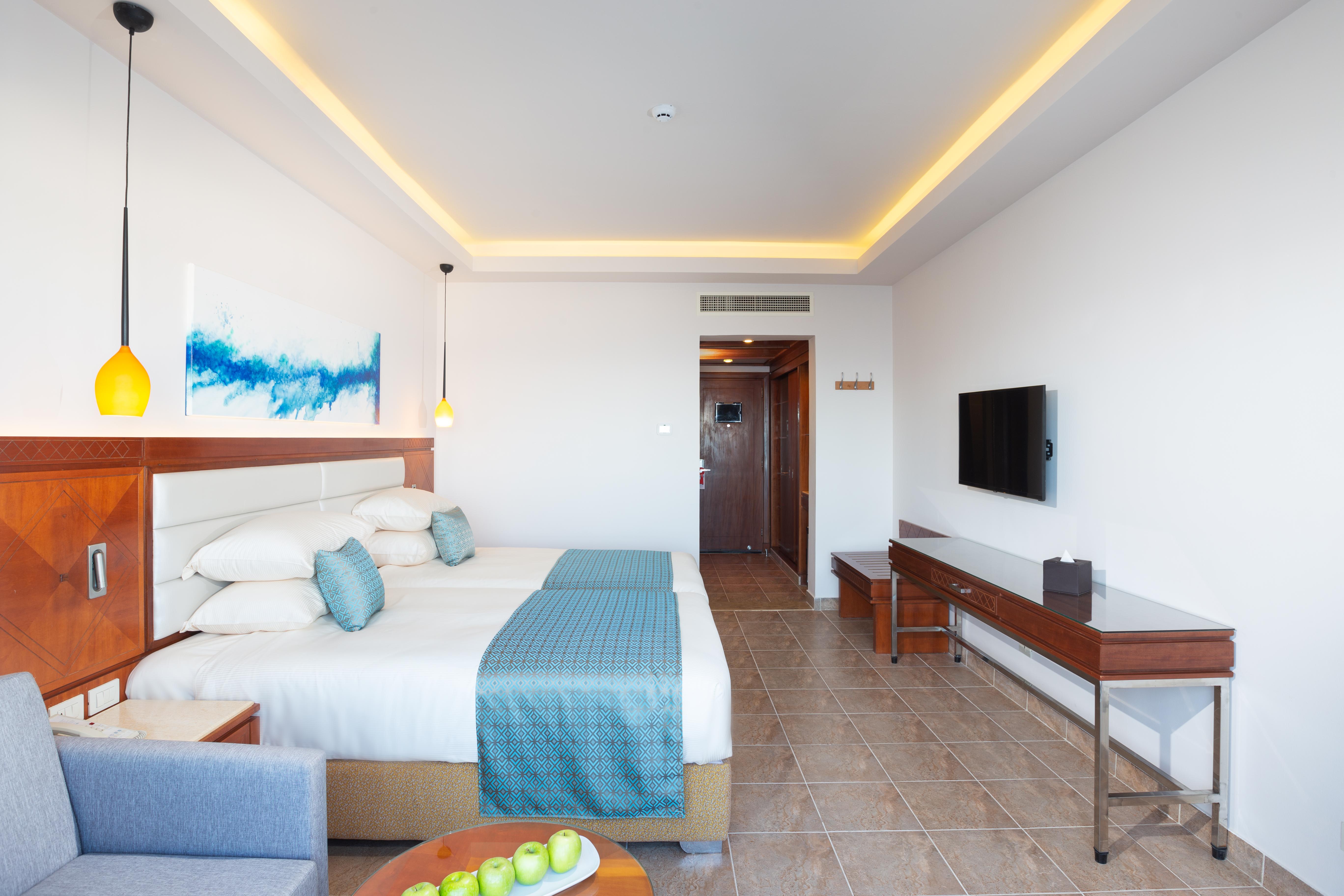 Pickalbatros Palace Sharm - "Aqua Park" Exterior foto The photo shows a modern hotel room featuring two queen-sized beds with white linens and a light blue throw. The room is well-lit, with a warm yellow glow from the ceiling lights. There is a painting on the wall above the beds, characterized by blue 