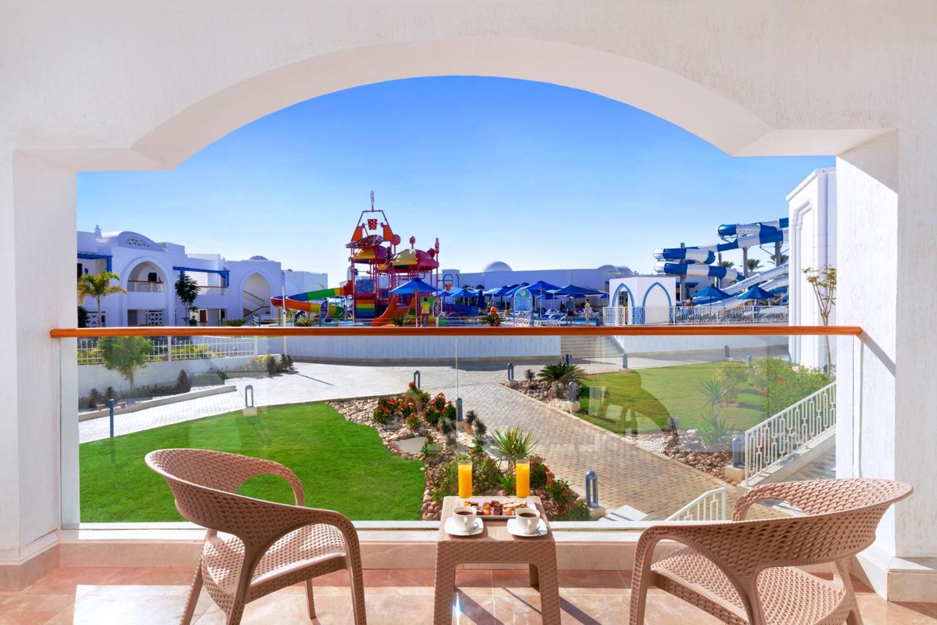Pickalbatros Palace Sharm - "Aqua Park" Exterior foto The photo shows a view from a balcony with two chairs and a small table set up for relaxation. In the foreground, there is a breakfast setup with orange juice and a pastry. Beyond the balcony, there is a vibrant, colorful water play area with slides 