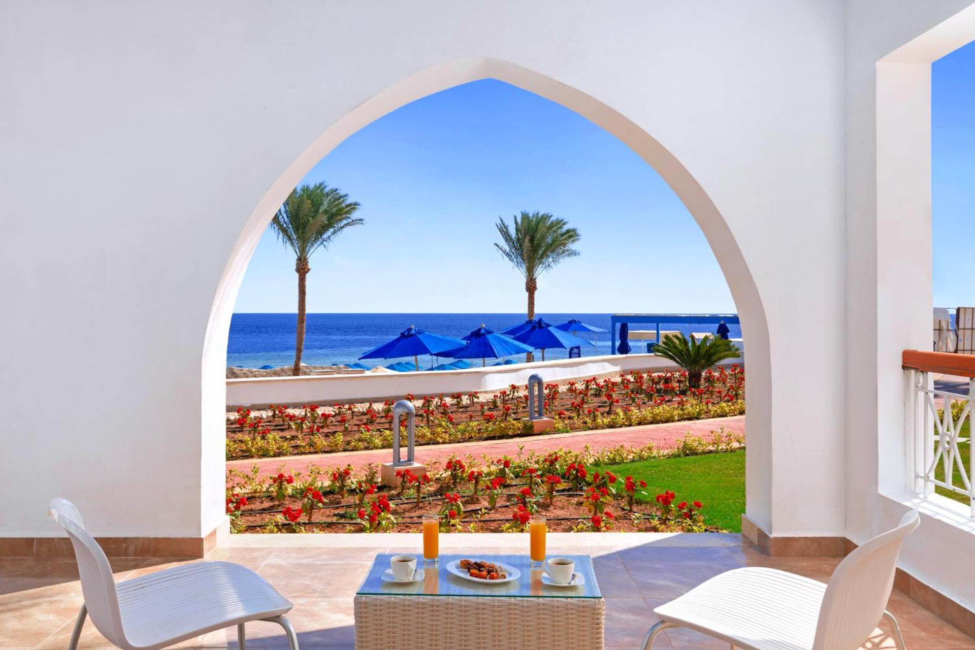 Pickalbatros Palace Sharm - "Aqua Park" Exterior foto The photo depicts a serene view through an architectural archway. In the foreground, there is a small table set with two glasses of orange juice and a plate of snacks. Surrounding the table are two white chairs. Beyond the arch, you can see a calm bl