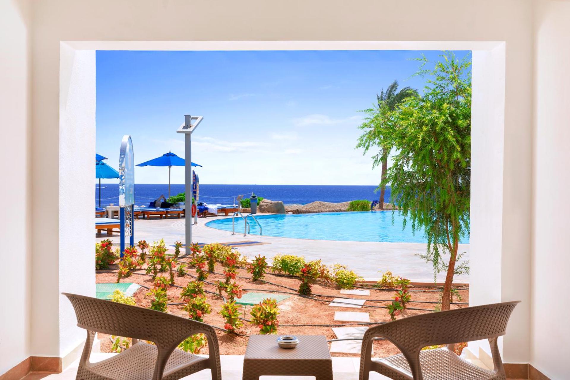 Pickalbatros Palace Sharm - "Aqua Park" Exterior foto The photo shows a serene view from inside a space looking out onto a beautiful scene. In the foreground, there are two comfortable chairs positioned around a small table, suggesting a cozy place to relax. Beyond the seating area, there is a landscape