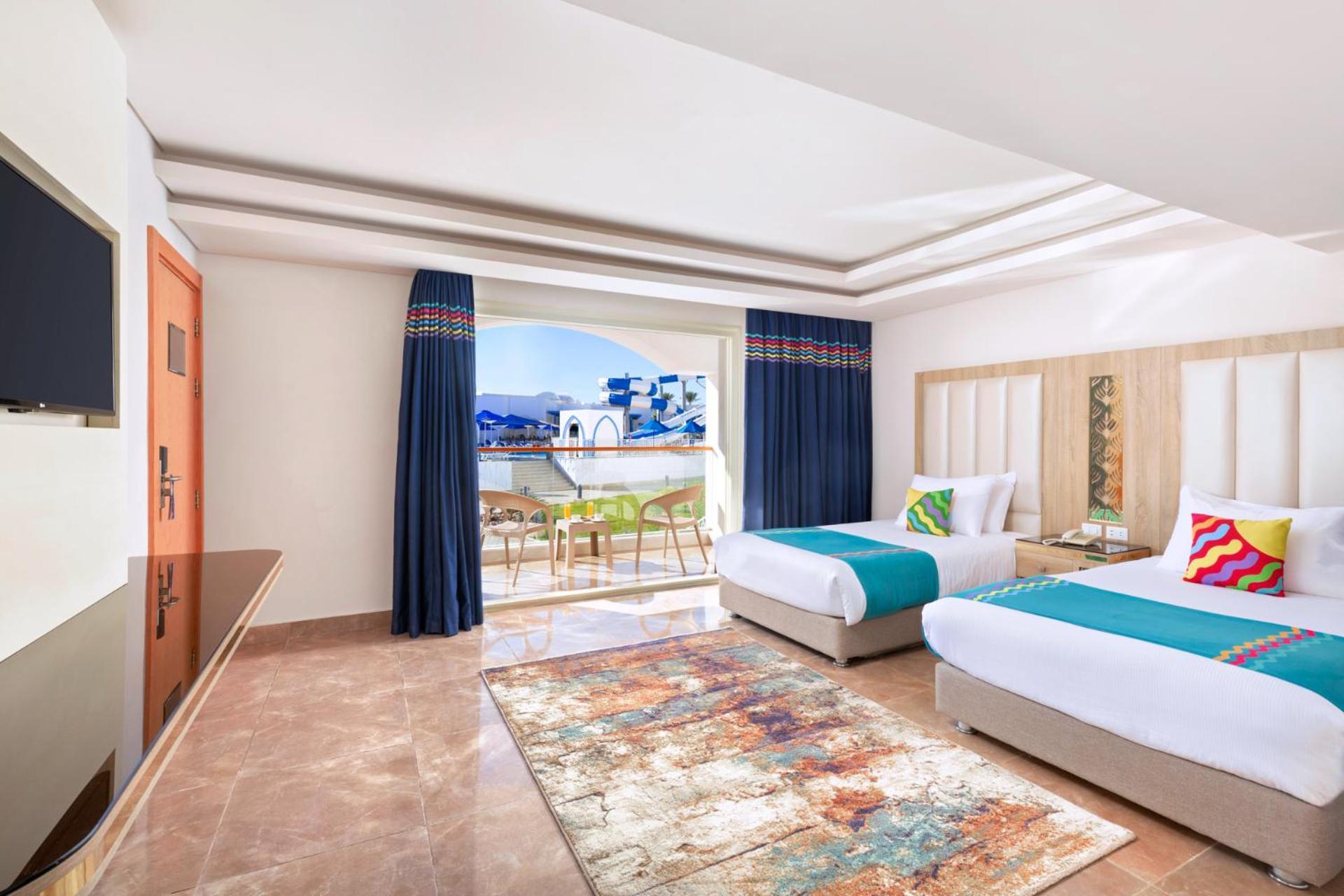 Pickalbatros Palace Sharm - "Aqua Park" Exterior foto The photo shows a well-decorated hotel room featuring two beds, each with colorful accent pillows. The walls are painted in a light color, and there is a large window with blue curtains that offers a view of an outdoor area. A small seating area with